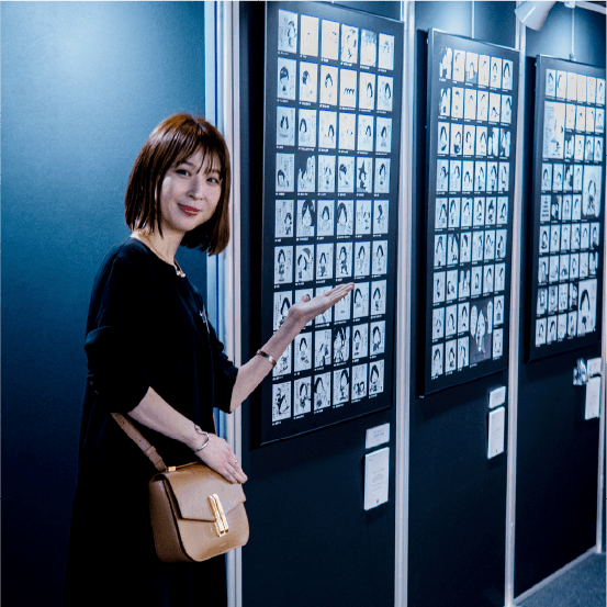 １コマ漫画のパネルの横で柔和な表情で立っているアッシー。左手でパネルを紹介している。